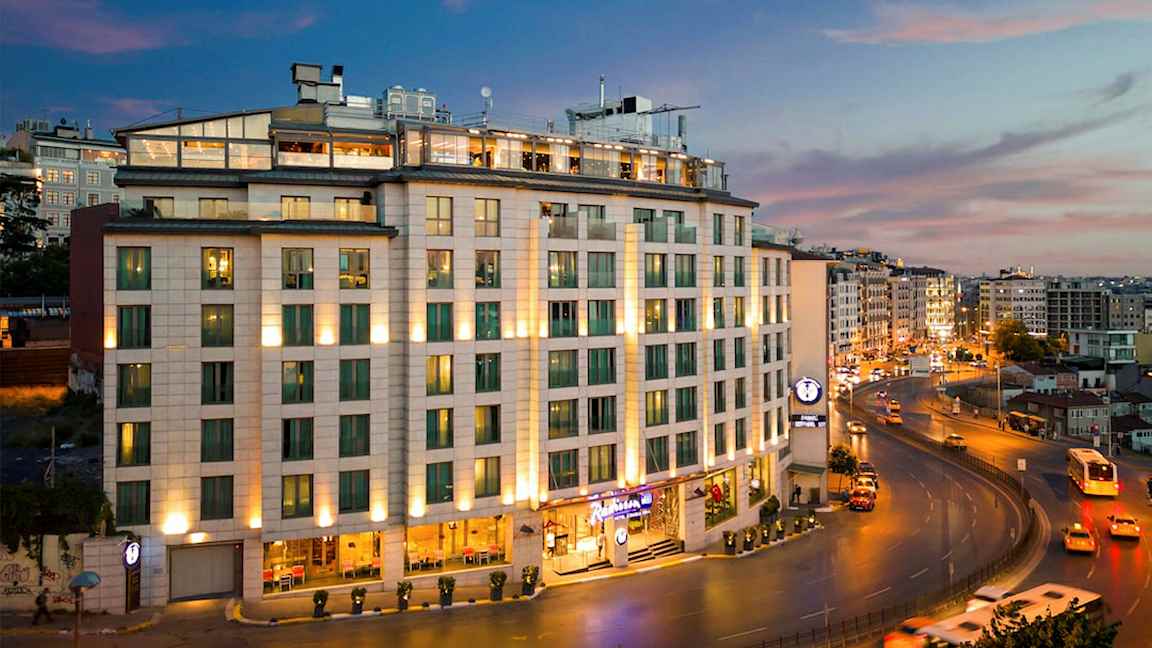 Nykyaikainen rakennus, jonka julkisivua valaisee ja jossa on suuret ikkunat. Se sijaitsee suuren tien varrella kaupunkialueella, ja ohi kulkee autoja ja linja-autoja. (Tekoälyn luoma teksti)