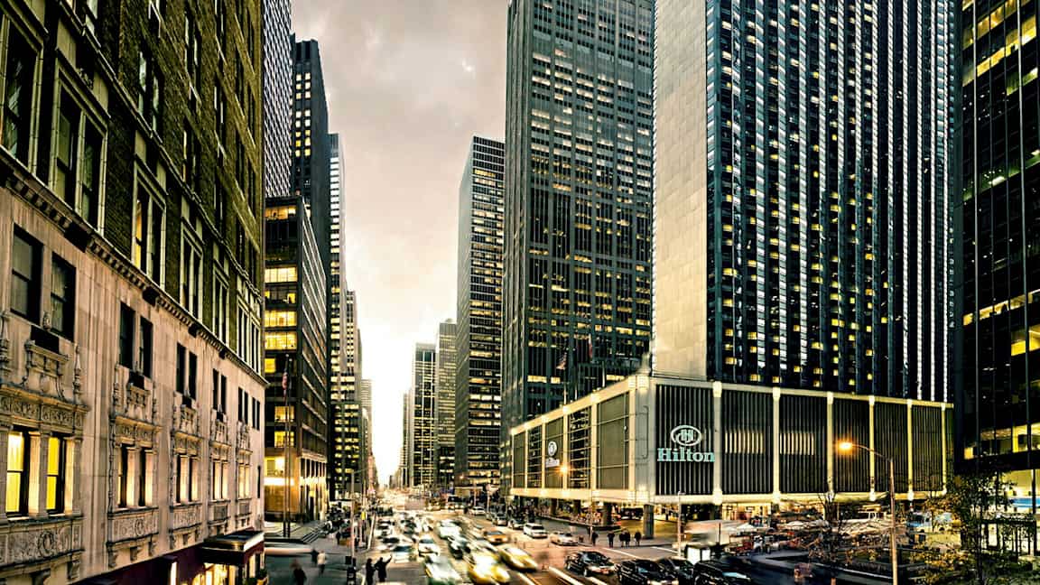 Kaupunki näkymä, jossa on korkeita pilvenpiirtäjiä, vilkasta liikennettä ja rakennuksia, joissa on lukuisia ikkunoita. Hilton-hotelli näkyy alareunassa. (Tekoälyn luoma teksti)