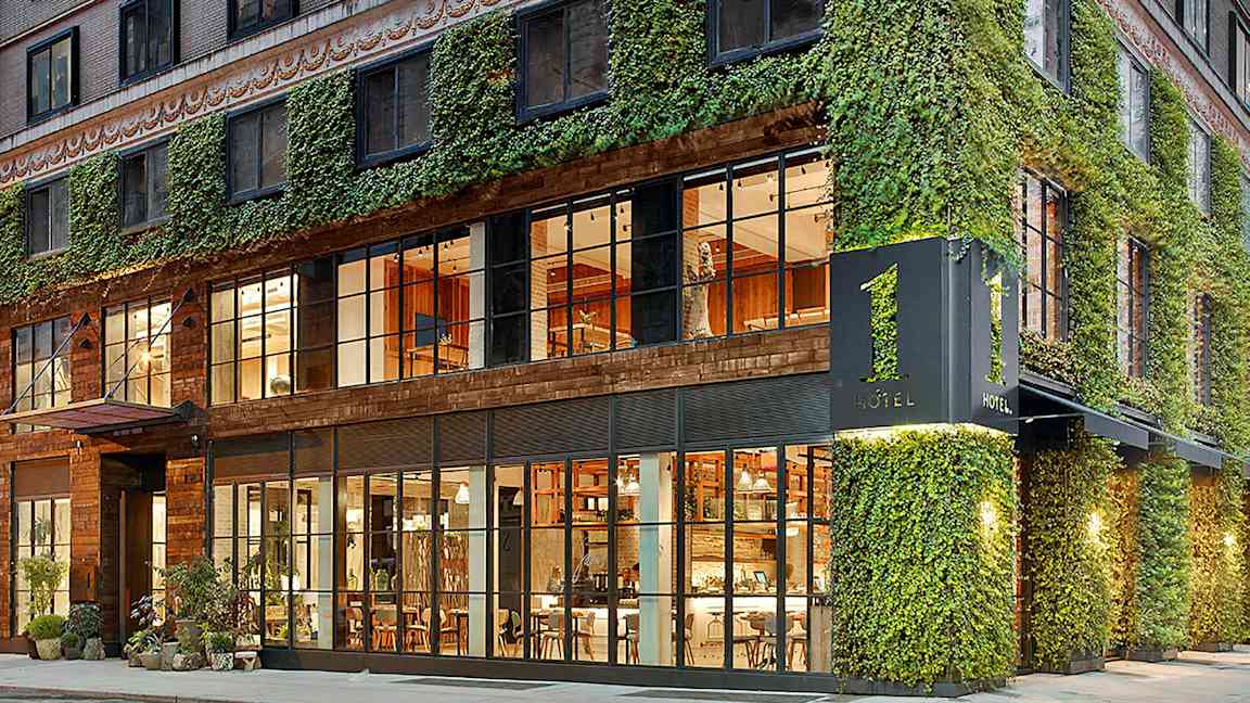 Hotellin julkisivu, jossa on muratin peittämä seinä ja suuret lasi-ikkunat, joista näkyy sisätilojen lämmin valaistus. (Tekoälyn luoma teksti)