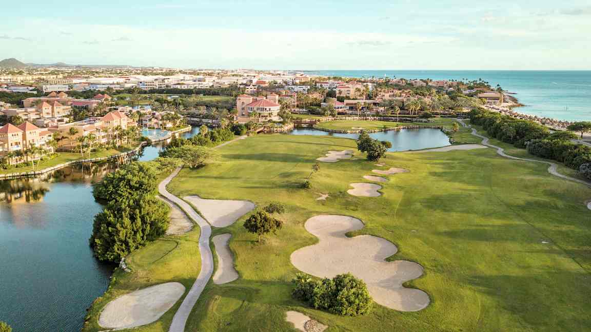 Golfkenttä, jossa on hiekkabunkkereita ja vihreitä nurmialueita lähellä vesiväylää ja rantaa. Taustalla rakennuksia. (Tekoälyn luoma teksti)