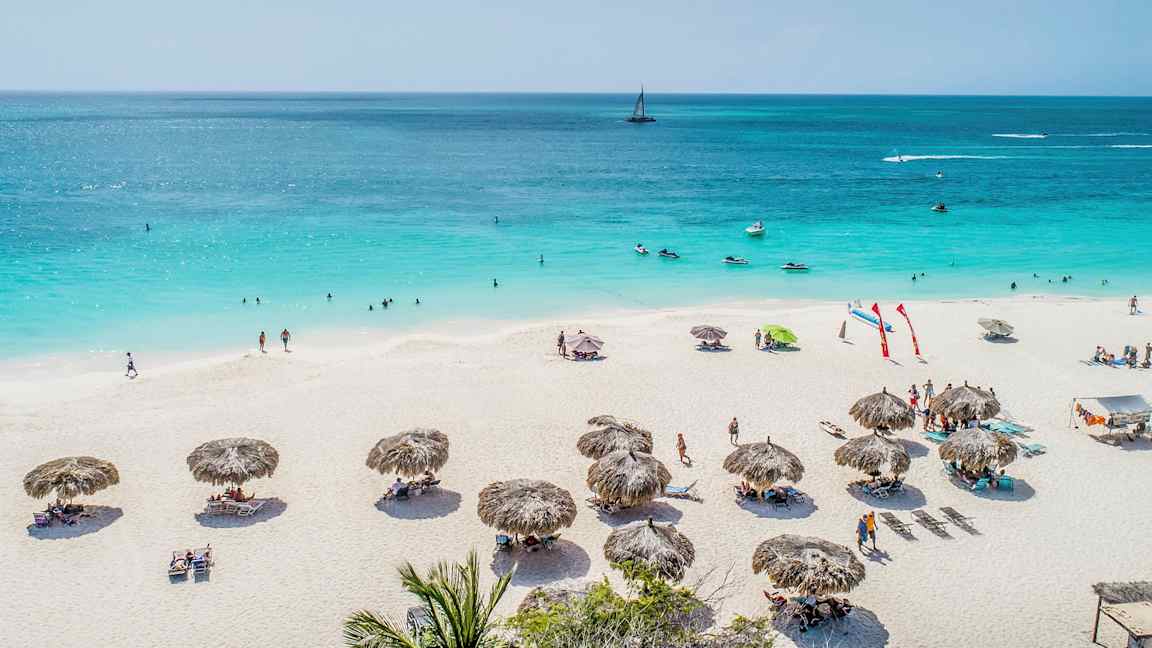 Hiekkaranta, jossa aurinkovarjoja ja ihmisiä, jotka ottavat aurinkoa ja uivat sinisessä merimaisemassa. (Tekoälyn luoma teksti)