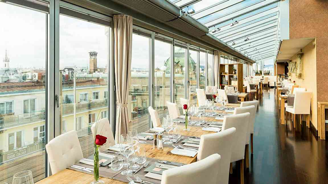 Valoisa ravintola, jossa on lasiseinät ja -katto, pöydät on katettu valkoisilla tuoleilla ja jokaiseen pöytään on asetettu ruusu. (Tekoälyn luoma teksti)