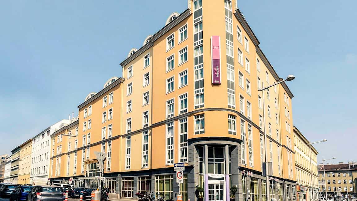 Vaaleankeltainen rakennus, jossa on useita ikkunoita, kyltti ja pyöreä kulmajulkiin. (Tekoälyn luoma teksti)