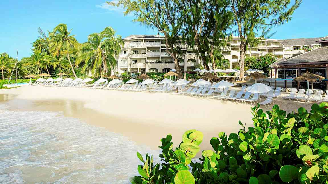 Ranta, jossa on aurinkotuoleja ja -varjoja lomakeskuksen edessä, ympärillä palmuja ja vehreyttä. (Tekoälyn luoma teksti)