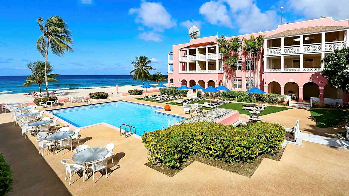 Kuva aurinkoisesta lomakeskuksesta, jossa on ulkouima-allas, ympäröiviä ulkoalueita ja vaaleanpunainen hotelli parvekkeella, meren rannalla. (Tekoälyn luoma teksti)