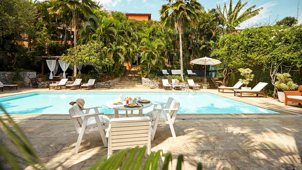 Ulkouima-allas, jota ympäröi kasvillisuus ja aurinkotuolit. Pöytä, jossa on ruokaa ja juomaa, seisoo altaan reunalla. (Tekoälyn luoma teksti)