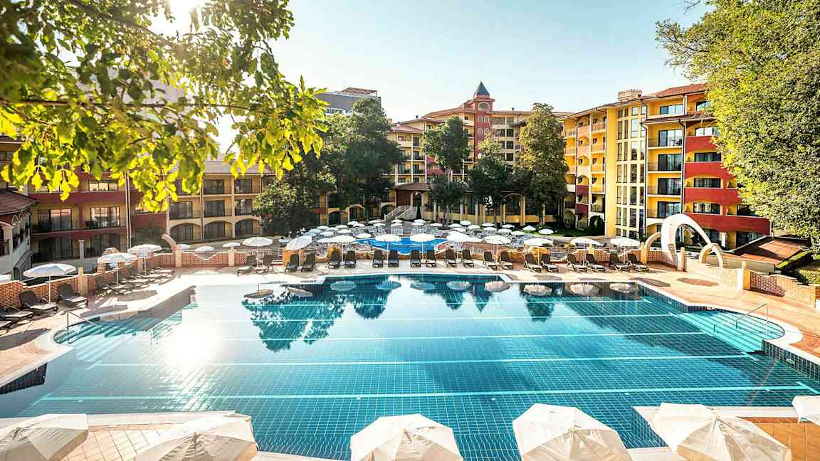 Ulkoallas, jota ympäröivät aurinkotuolit ja aurinkovarjot, taustalla monivärisiä rakennuksia ja puita. (Tekoälyn luoma teksti)