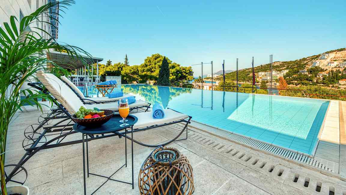 Näkymä infinity-uima-altaan suuntaan, jossa on aurinkotuoleja, pöytiä hedelmien ja juomien kanssa vieressä sekä vihreä ympäristö ja taivas taustalla. (Tekoälyn luoma teksti)