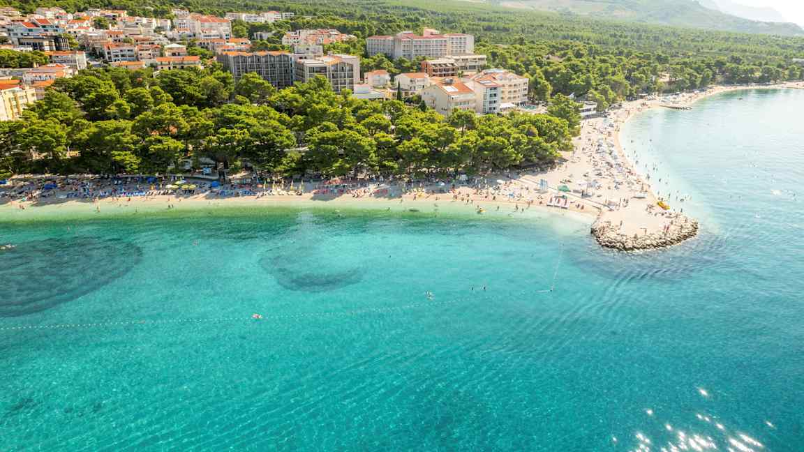 Ilmakuva rannasta, jossa kirkas sininen vesi, aurinkotuoleja ja tiivis asutus metsän ja vuorten ympäröimänä. (Tekoälyn luoma teksti)