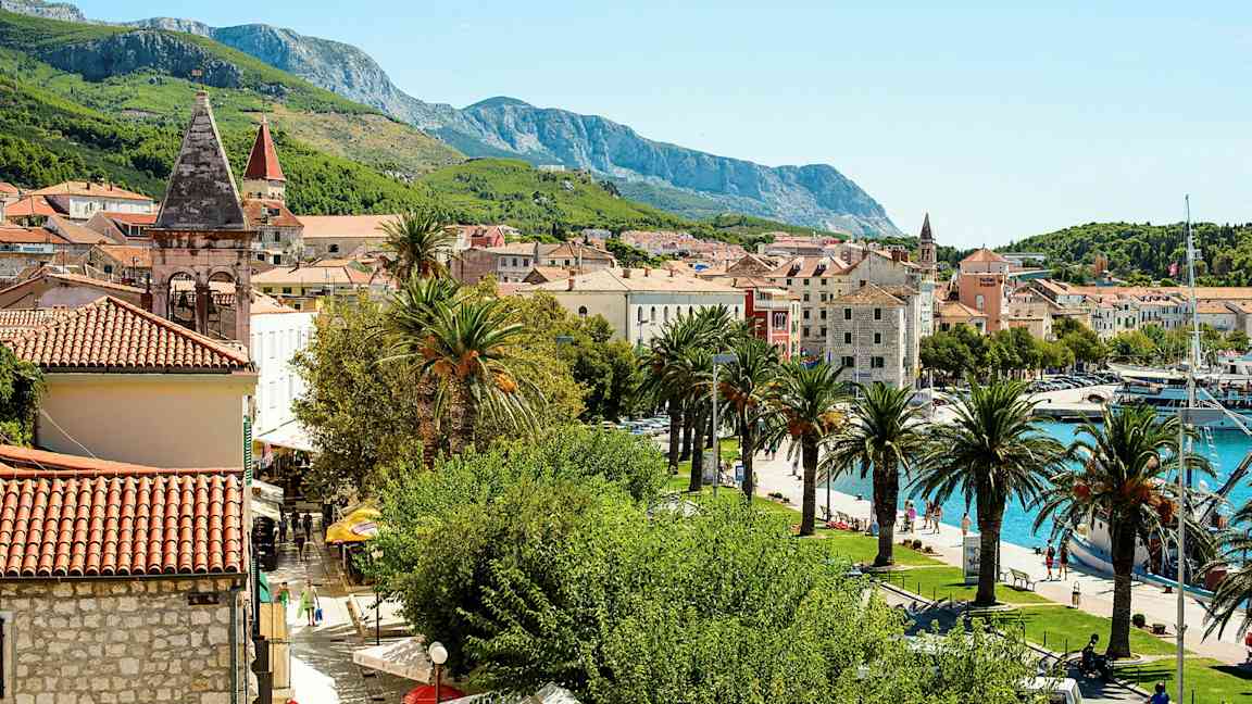 Kuva esittää viehättävää välimerellistä kaupunkia, jossa on punatiilikattoja, kirkontorneja, palmuja ja vehreitä vuoria taustalla. Kuvassa näkyy myös venesatama. (Tekoälyn luoma teksti)