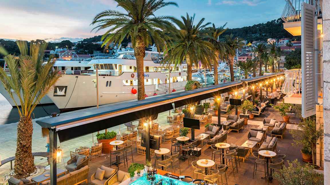 Näkymä ulkoilmaravintolasta satamaan, jossa on palmupuita ja suuria veneitä. (Tekoälyn luoma teksti)