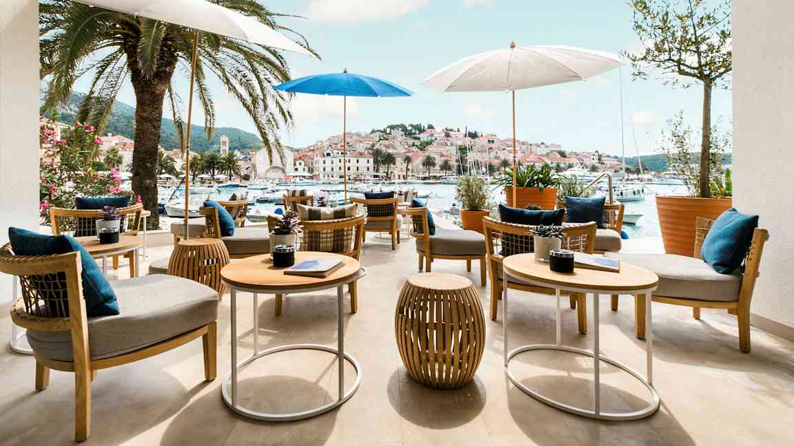 Ulkoterassi, jossa pöytiä ja tuoleja varjojen alla, ympärillä palmuja ja näkymä viehättävään satamaan ja kaupunkiin. (Tekoälyn luoma teksti)