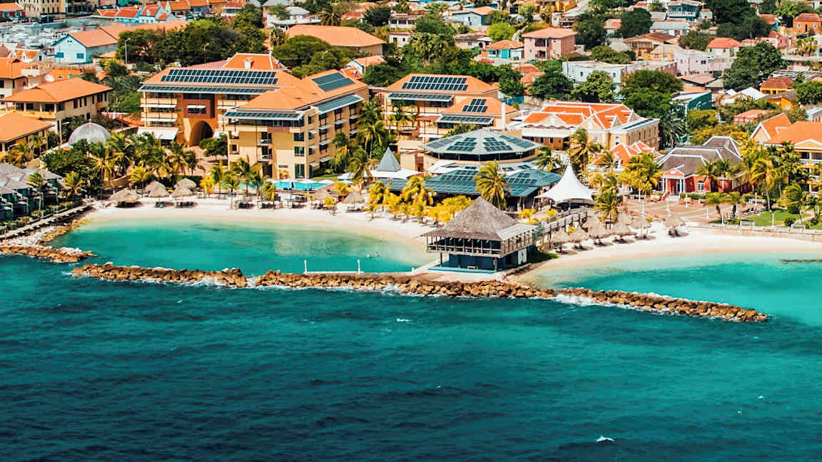 Ilmakuva rannikkokaupungista, jossa turkoosia vettä, hiekkaranta ja rakennuksia punaisilla katoilla ja aurinkopaneeleilla. (Tekoälyn luoma teksti)