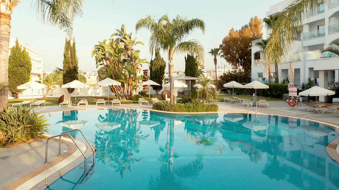 Ulkouima-allas, jota ympäröivät aurinkotuolit ja aurinkovarjot, palmuja ja rakennuksia taustalla. (Tekoälyn luoma teksti)