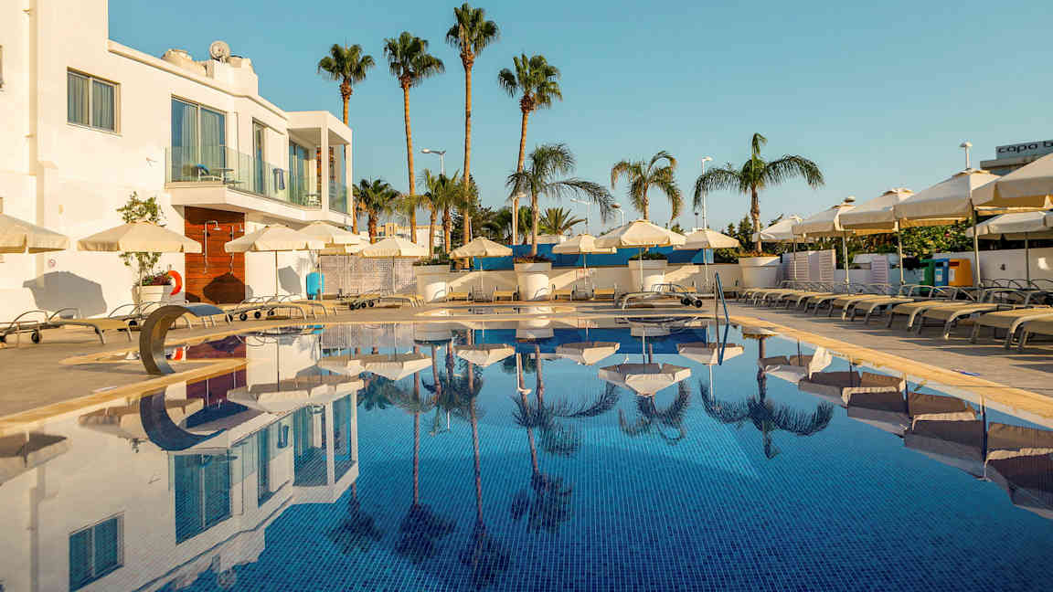 Kuva ulkouima-altaasta, jonka ympärillä on aurinkotuoleja ja aurinkovarjoja, taustalla palmuja ja valkoinen rakennus. (Tekoälyn luoma teksti)