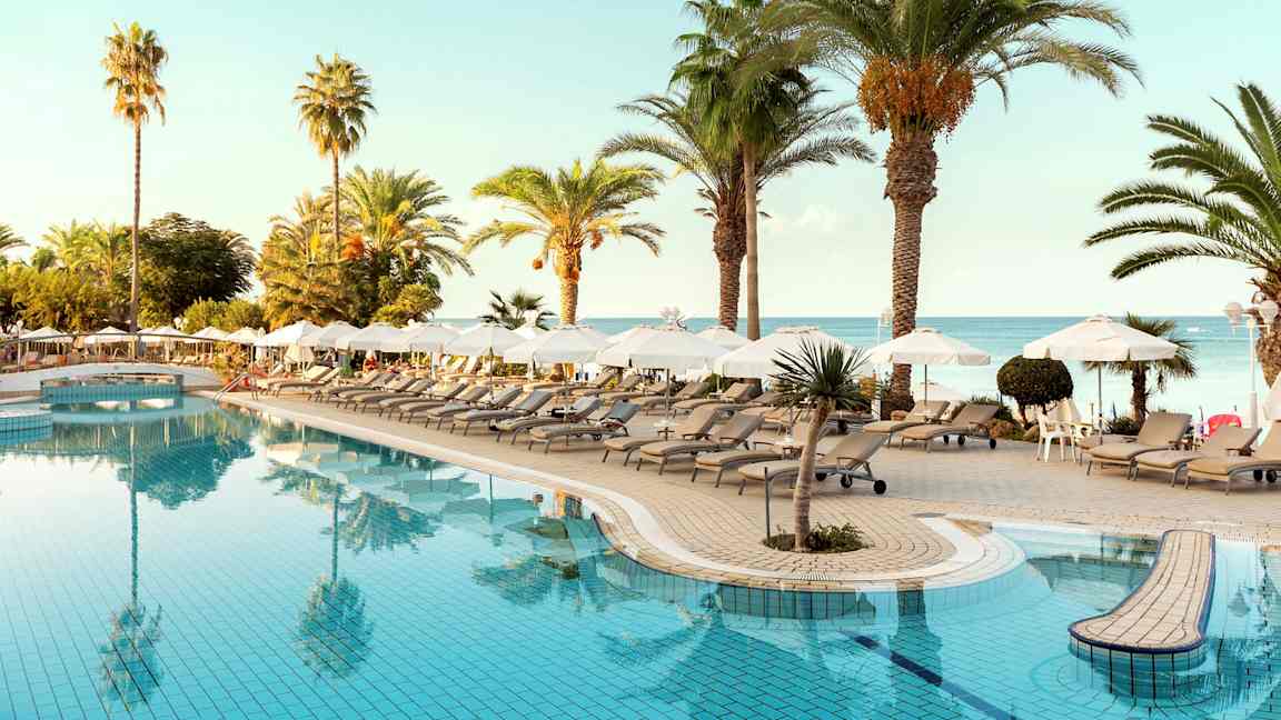 Uima-allasalue, jossa aurinkotuoleja, aurinkovarjoja ja palmuja lähellä merta. (Tekoälyn luoma teksti)