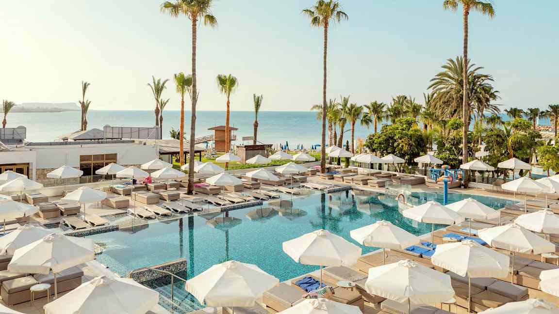 Ulkoallasalue, jossa aurinkotuoleja ja aurinkovarjoja, palmuja ympärillä ja lähellä merta. (Tekoälyn luoma teksti)