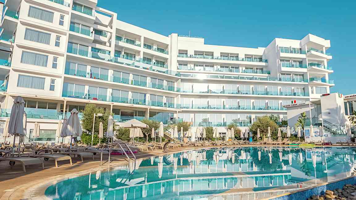 Valkoinen rakennus, jossa on useita kerroksia ja parvekkeita, uima-altaan edessä, jota ympäröivät aurinkotuolit ja aurinkovarjot. (Tekoälyn luoma teksti)