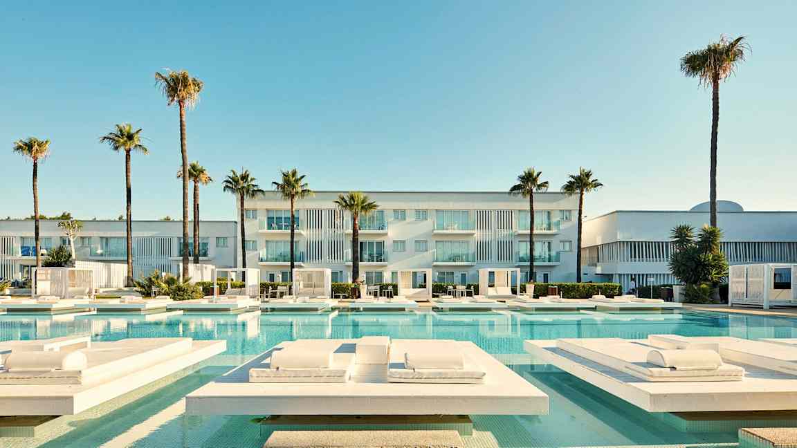 Moderni rakennus, jossa parvekkeet, ympäröity palmilla ja uima-altaalla, jossa on aurinkotuoleja. (Tekoälyn luoma teksti)