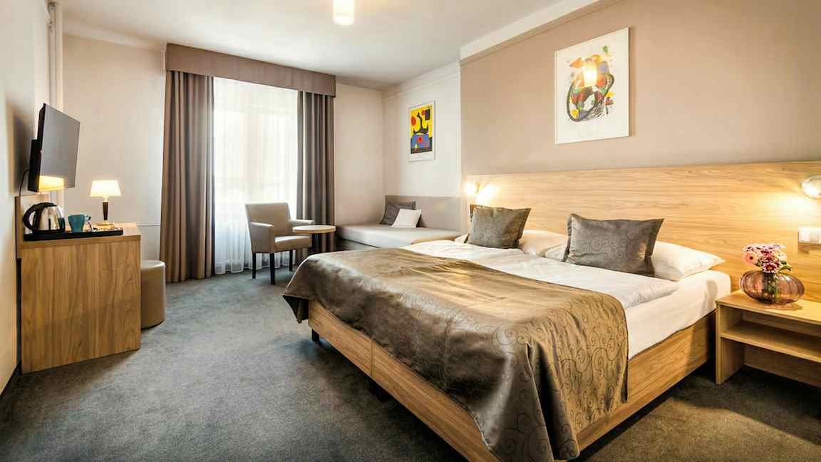 Hotellihuone, jossa on suuri parisänky, pöytä, kaksi tuolia ja seinälle asennettu televisio. (Tekoälyn luoma teksti)