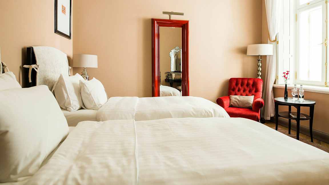 Hotellihuone, jossa kaksi sänkyä, punainen nojatuoli, pieni pöytä ja suuri ikkuna vaaleilla verhoilla. (Tekoälyn luoma teksti)