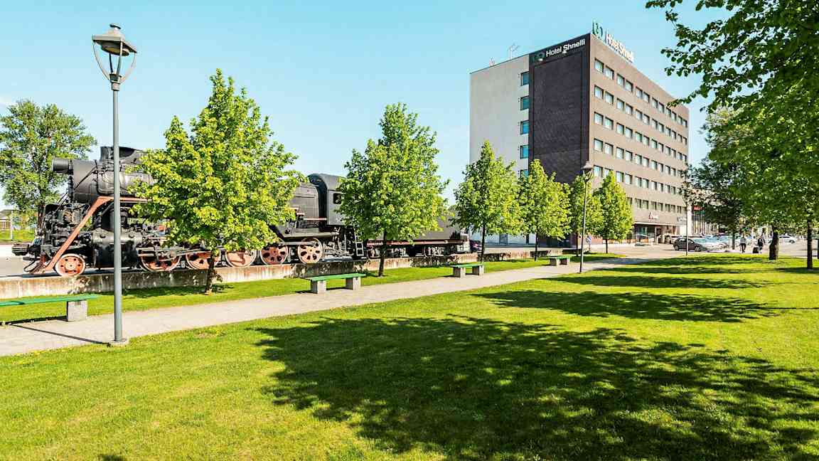 Vanha höyryveturi seisoo ulkona modernin hotellin vieressä vehreiden puiden keskellä. (Tekoälyn luoma teksti)