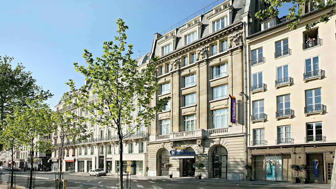 Katu, jolla on rakennuksia, puita ja ihmisiä aurinkoisessa säässä. (Tekoälyn luoma teksti)