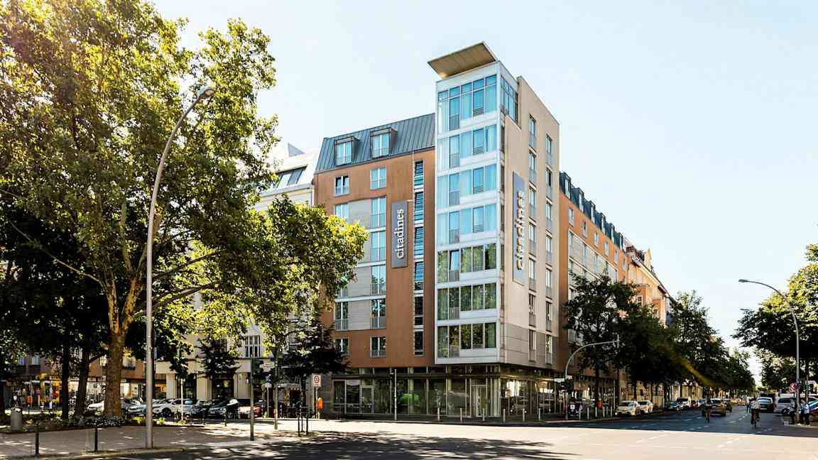 Moderni hotelli lasiseinällä, ympärillä puita ja kaupunkimaisemaa. (Tekoälyn luoma teksti)