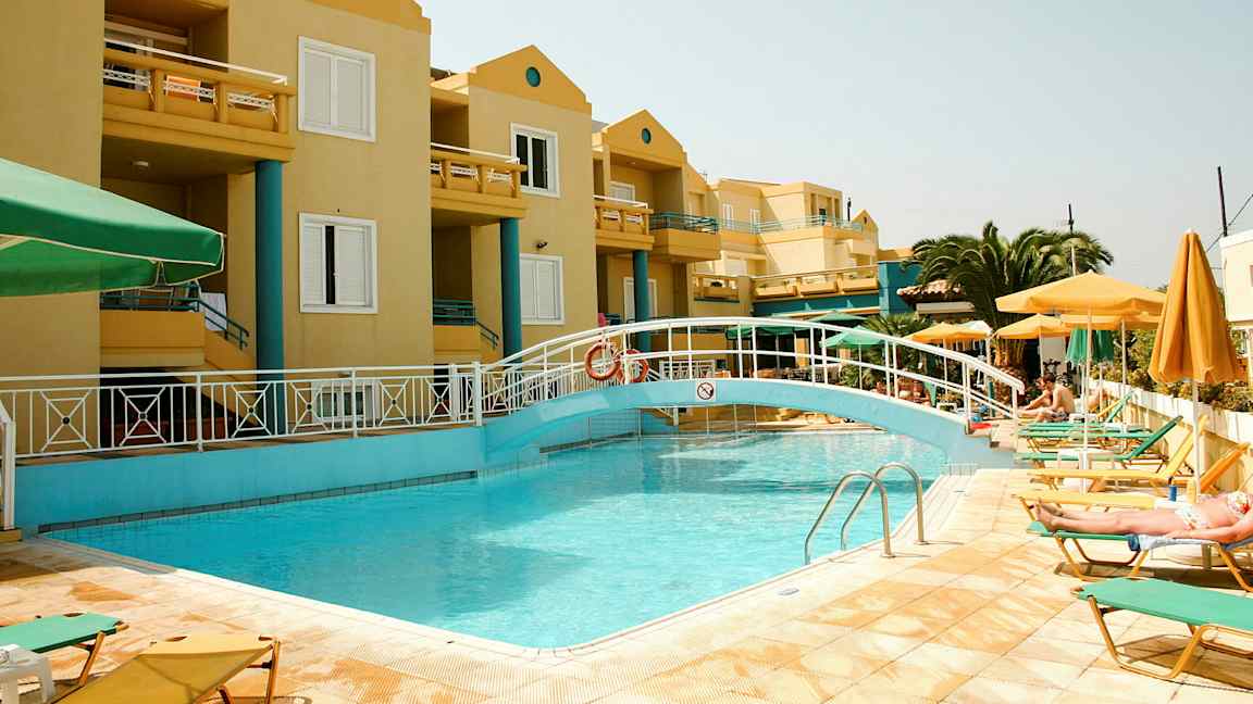 Uima-allas, jossa on silta keltaisen rakennuksen edessä, ympärillä on aurinkotuoleja ja varjoja. (Tekoälyn luoma teksti)