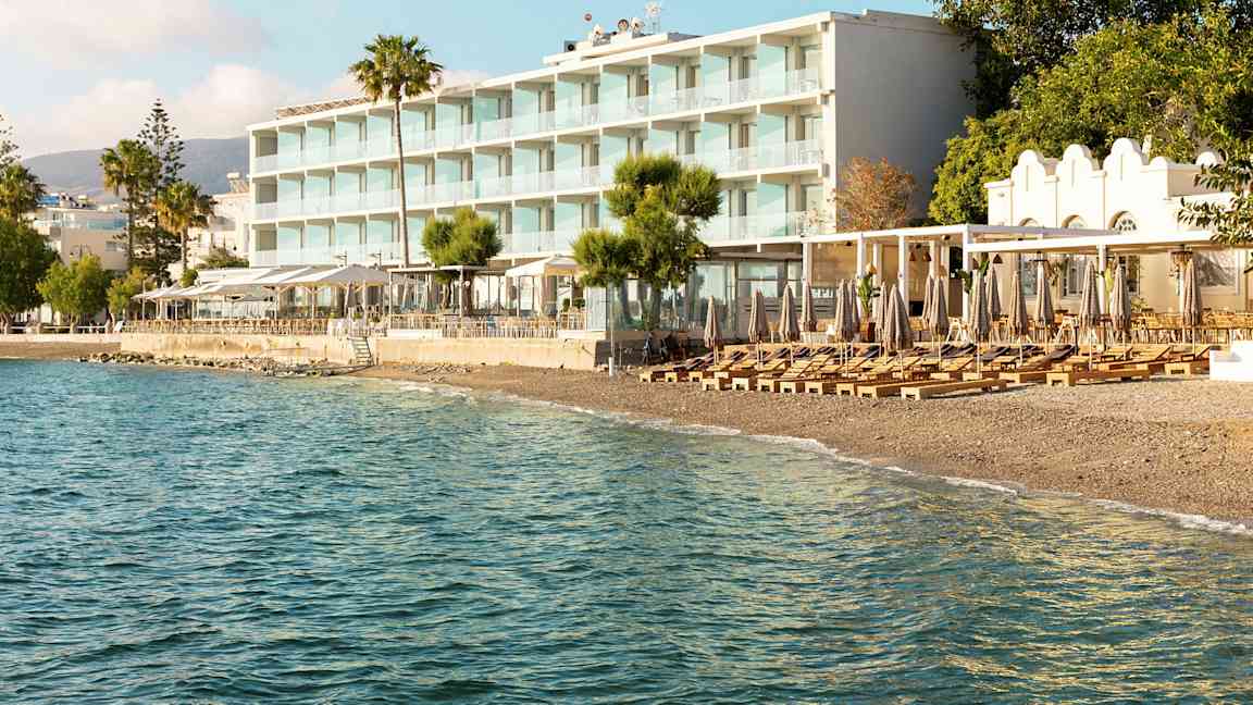 Näkymä rantahotelliin, jossa on aurinkotuoleja ja aurinkovarjoja rannalla ja meri etualalla. (Tekoälyn luoma teksti)