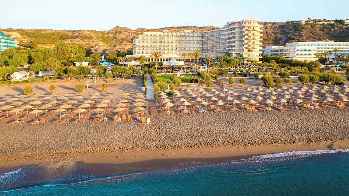 Ranta aurinkovarjoilla ja aurinkotuoleilla, taustalla hotelli ja vihreitä alueita. (Tekoälyn luoma teksti)