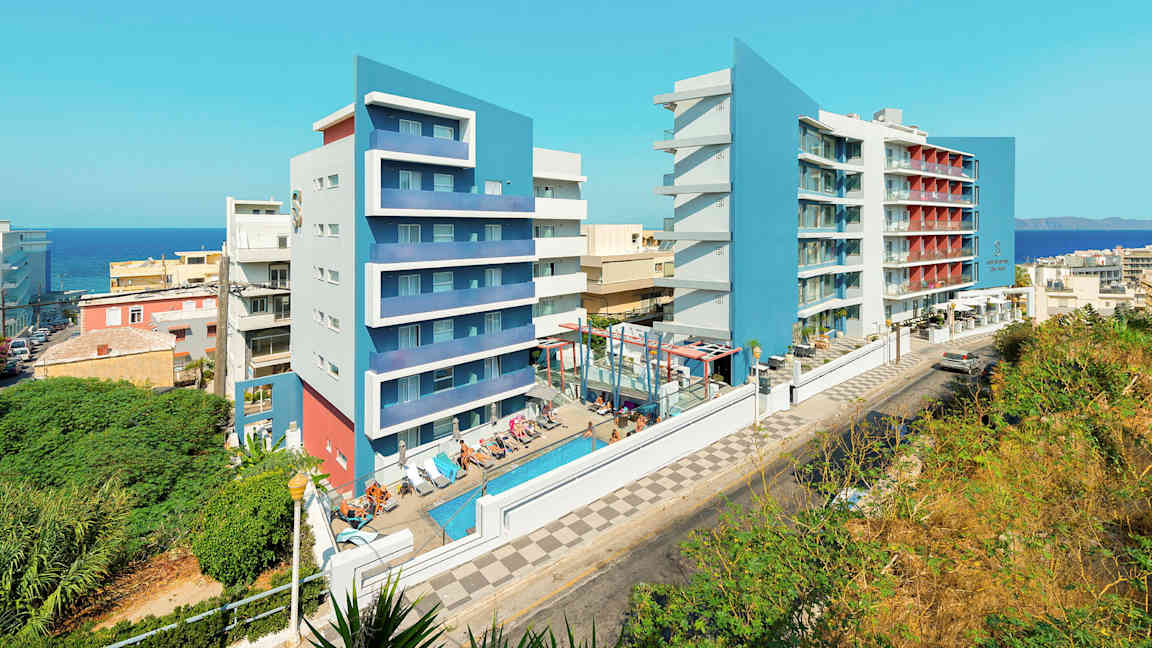 Kuva kahdesta modernista hotellirakennuksesta, joissa on siniset julkisivut, sekä uima-allas ja aurinkotuoleja etualalla. (Tekoälyn luoma teksti)