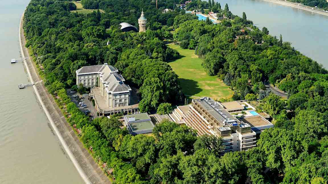 Ilmakuva vehreästä saaresta, jossa on rakennuksia, nurmikkoalueita ja puita joen ympäröimänä. (Tekoälyn luoma teksti)