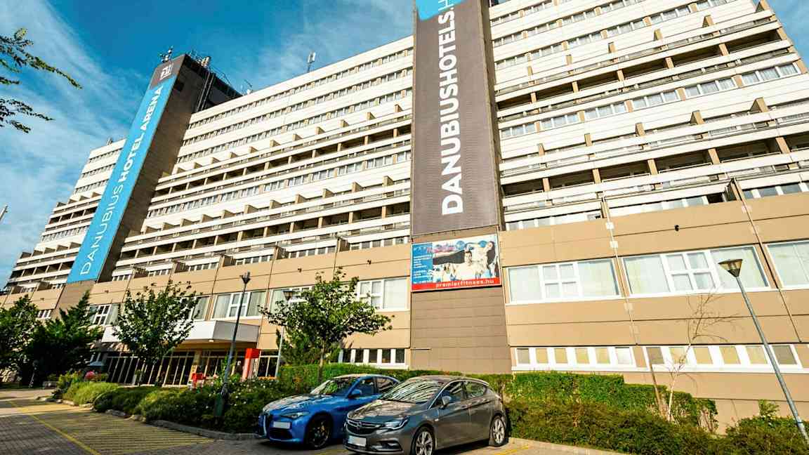 Suuri hotellirakennus, jossa on useita kerroksia ja näkyvässä kyltissä lukee ”Danubius Hotel Arena”. Rakennuksen edessä on pysäköityjä autoja ja joitakin pieniä puita. (Tekoälyn luoma teksti)