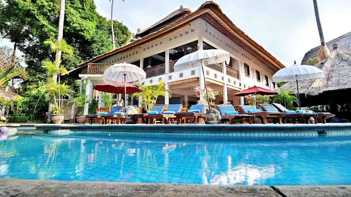 Iso huvila olkikatolla ja uima-allas edessä, ympärillä aurinkotuoleja ja varjoja. (Tekoälyn luoma teksti)