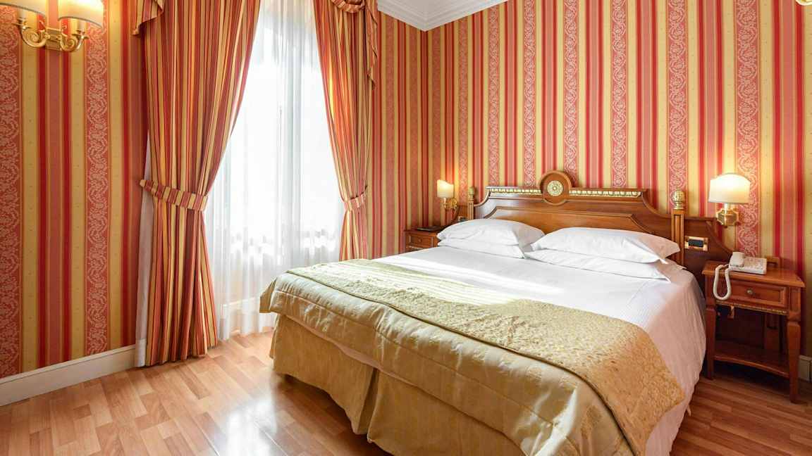 Makuuhuone, jossa on raidalliset punaiset tapetit, parivuode valkoisilla vuodevaatteilla ja kullankeltainen peite sekä puupöydät sivuilla. (Tekoälyn luoma teksti)