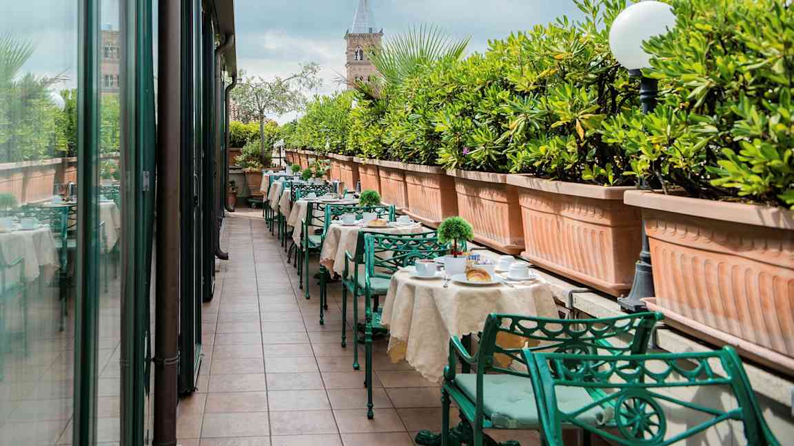 Ulkoterassi, jossa on katetut pöydät ja vihreät metallituolit, reunustettu suurilla viherkasveilla täytetyillä istutuslaatikoilla. (Tekoälyn luoma teksti)