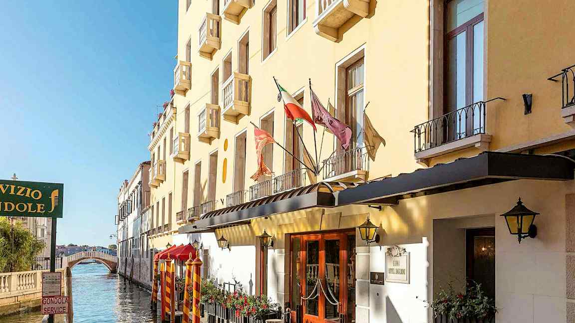 Keltainen rakennus, jossa on parvekkeita kanavan varrella, ja taustalla kävelysilta. (Tekoälyn luoma teksti)