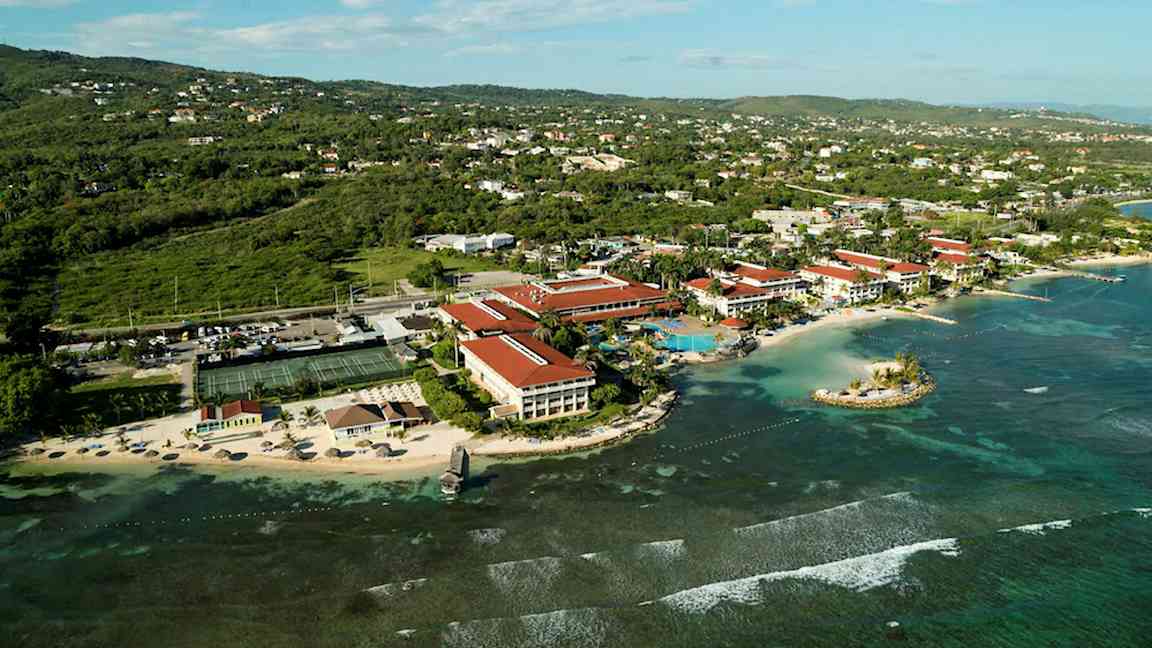 Ilmakuva rannikon lomakohteesta, jossa on punakattoisia rakennuksia, ranta, meri ja ympäröivä kasvillisuus. (Tekoälyn luoma teksti)