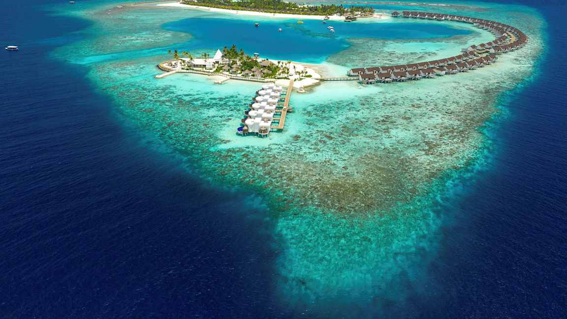 Ilmakuva trooppisesta saaresta, jossa veden yläpuolella olevia bungaloweja ja sininen laguuni, ympäröitynä koralliriutalla. (Tekoälyn luoma teksti)