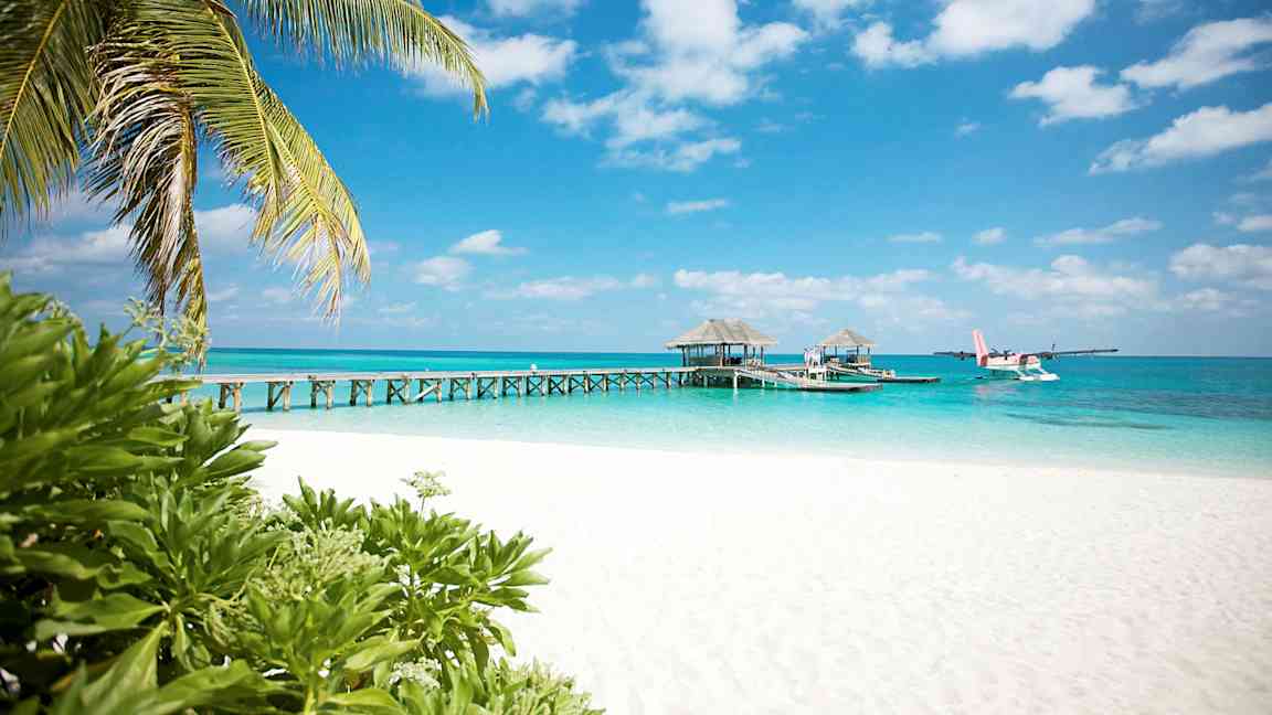 Valkoinen hiekkaranta, palmuja ja laituri, joka johtaa veden ylle resort-rakennuksiin. Vesitaso näkyy laiturin vieressä. (Tekoälyn luoma teksti)