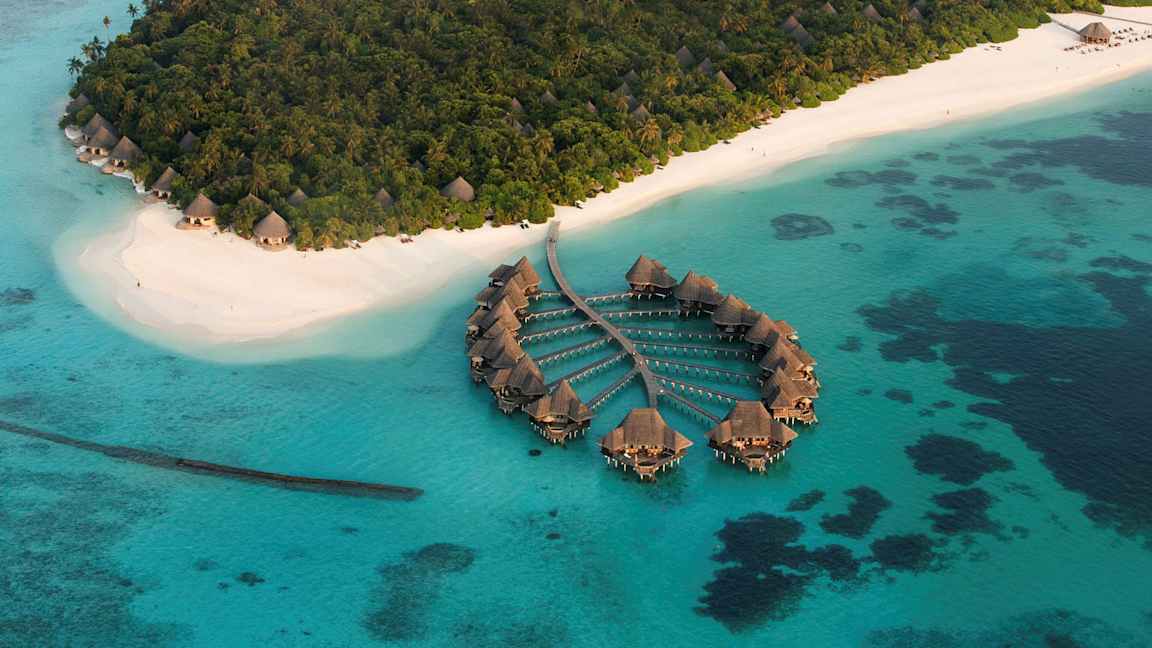 Ilmakuva trooppisesta saaresta, jossa on valkoiset hiekkarannat ja veden ylle rakennettuja huviloita. (Tekoälyn luoma teksti)