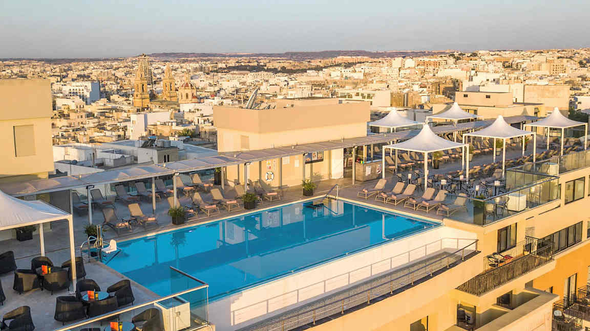 Kuva kattoterassista, jossa on uima-allas, aurinkotuoleja ja taustalla näkyvä kaupunkinäkymä. (Tekoälyn luoma teksti)