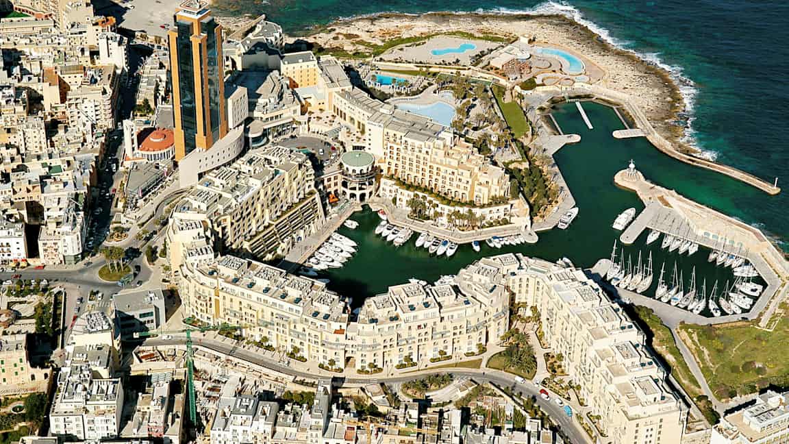 Ilmakuva rannikkokaupungista, jossa on korkeita rakennuksia, venesatama ja taustalla meri. (Tekoälyn luoma teksti)