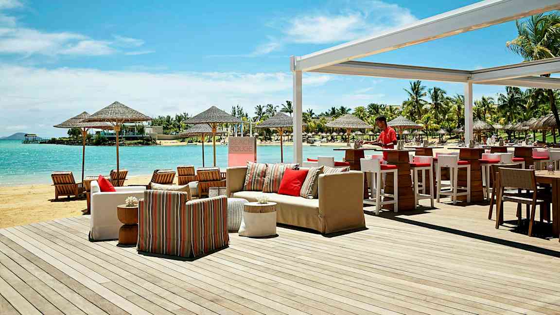 Ulkotarjoilualue rannalla, jossa on baari ja istumapaikkoja, sekä näkymä merelle ja ympäröiviin palmuihin. (Tekoälyn luoma teksti)