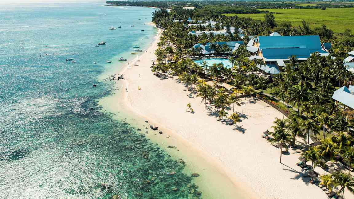 Näköala rannikolle, jolla leveä hiekkaranta, turkoosi meri ja uima-allasalue palmuineen sekä hotellirakennuksineen. (Tekoälyn luoma teksti)