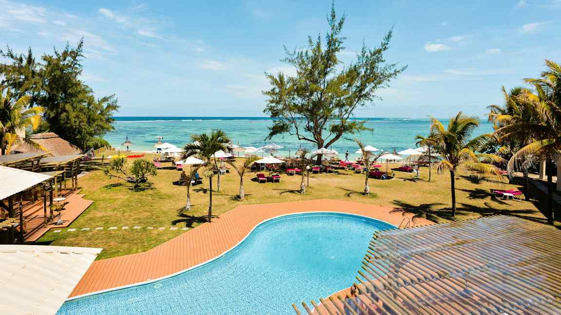 Uima-allas, jossa on turkoosia vettä, nurmikon, puiden, aurinkotuolien ja varjojen edessä rannan tuntumassa. (Tekoälyn luoma teksti)