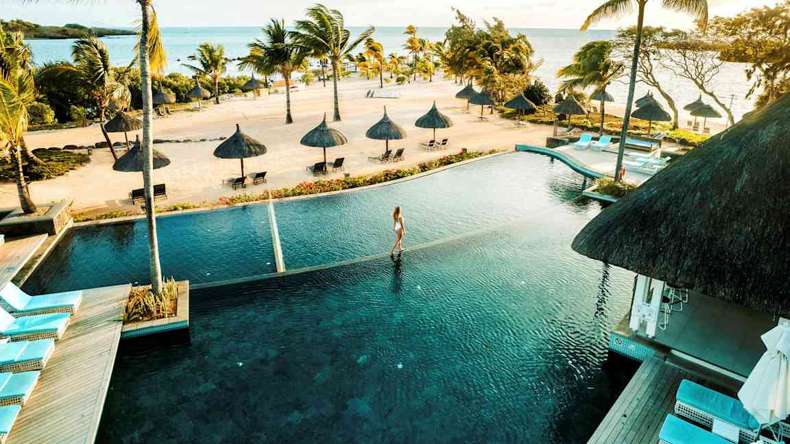 Trooppinen ranta, jolla on palmuja ja aurinkovarjoja meren äärellä, edessä infinity-allas. (Tekoälyn luoma teksti)