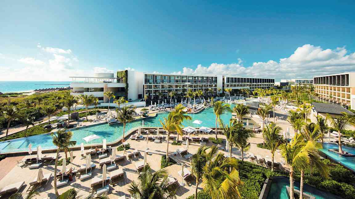 Nykyaikainen hotellialue, jossa on useita uima-altaita, vehreää kasvillisuutta ja palmuja, sijaitsee rannikolla ja tarjoaa näkymän merelle kirkkaan sinisen taivaan alla. (Tekoälyn luoma teksti)