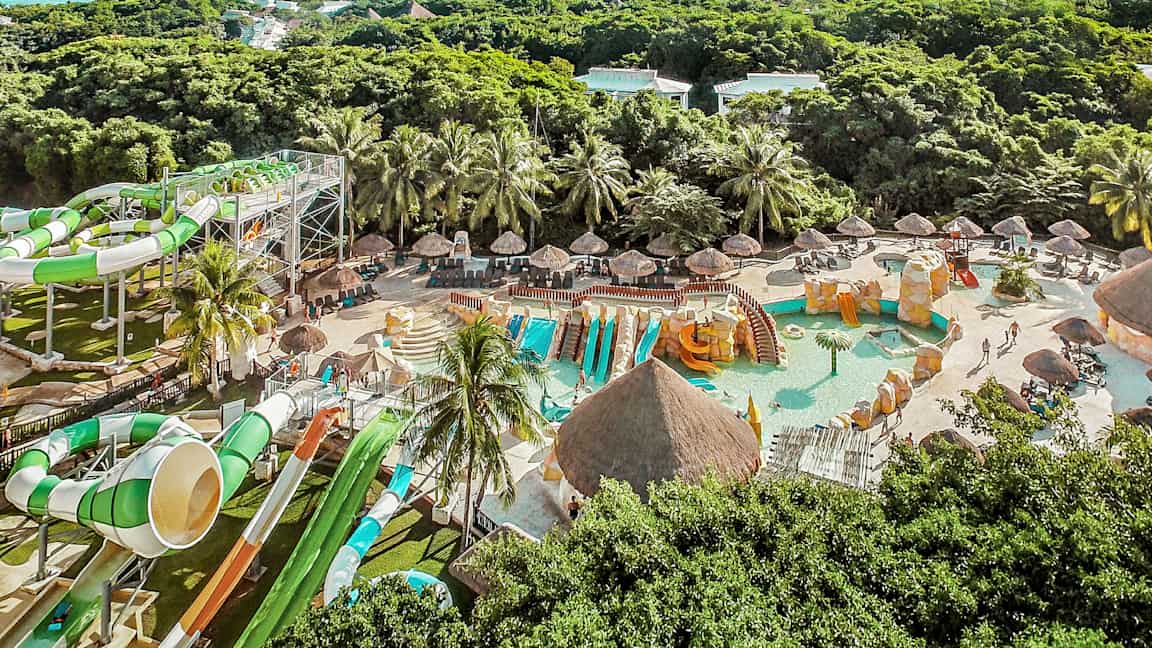 Kuva vesipuistosta, jossa on vesiliukumäkiä, uima-altaita ja vehreyttä taustalla. (Tekoälyn luoma teksti)